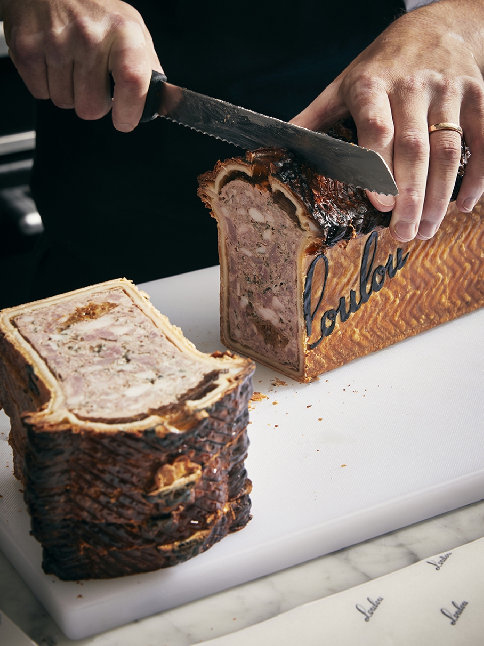 A person slices into a rectangular meat pie with a serrated knife on a white cutting board, showcasing the pie's layers and rich filling.
