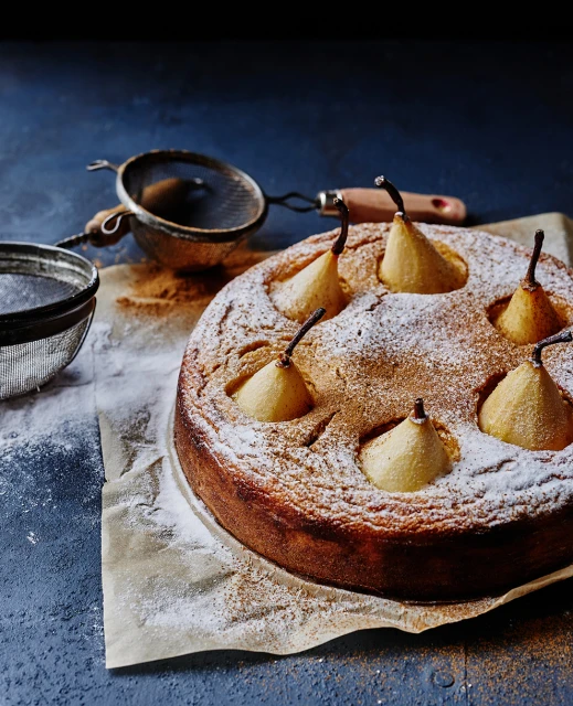 Overhead shot of a baked pear cake dusted with powdered sugar on parchment paper, next to metal sieves on a dark blue surface.
