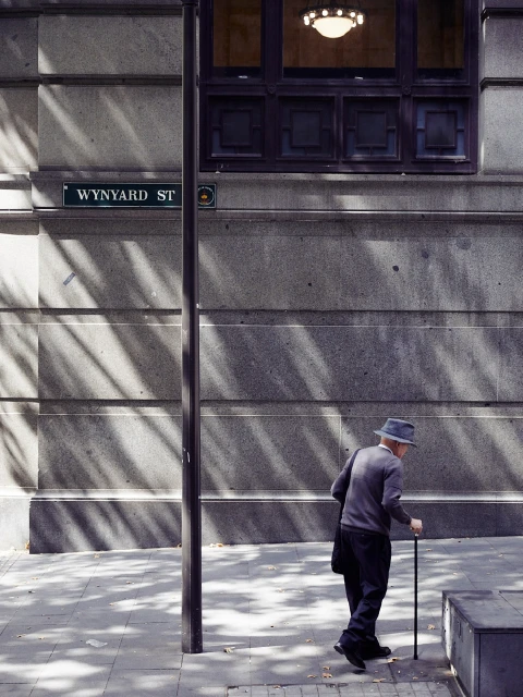 Elderly man with hat walks with cane on urban sidewalk with sunlight, shadow play and architectural details on city street.