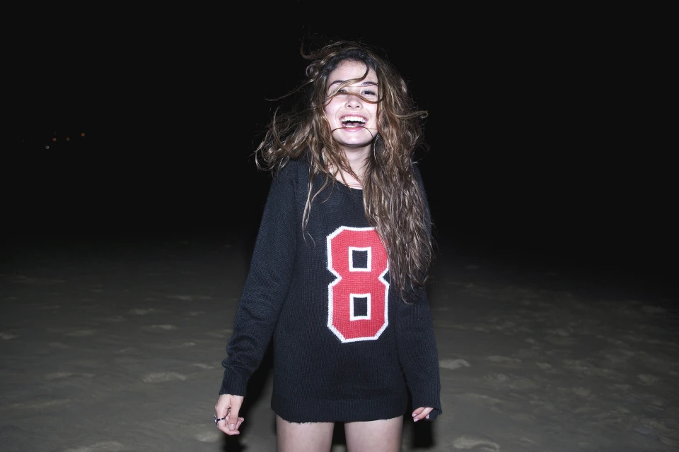 Young woman in black sweater with red number 8 stands on beach at night; the wind blows through her brown hair.