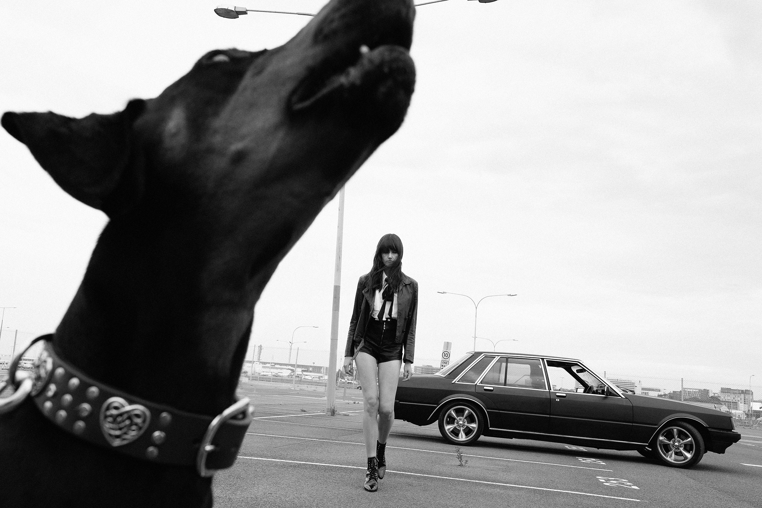 Black and white fashion shot of woman in leather jacket walking with a dog in a parking lot next to a vintage sedan car.
