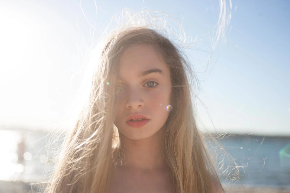 Portrait of a young girl with long blonde hair, standing outdoors with bright sunlight, ocean, and sky visible in the soft-focus background.