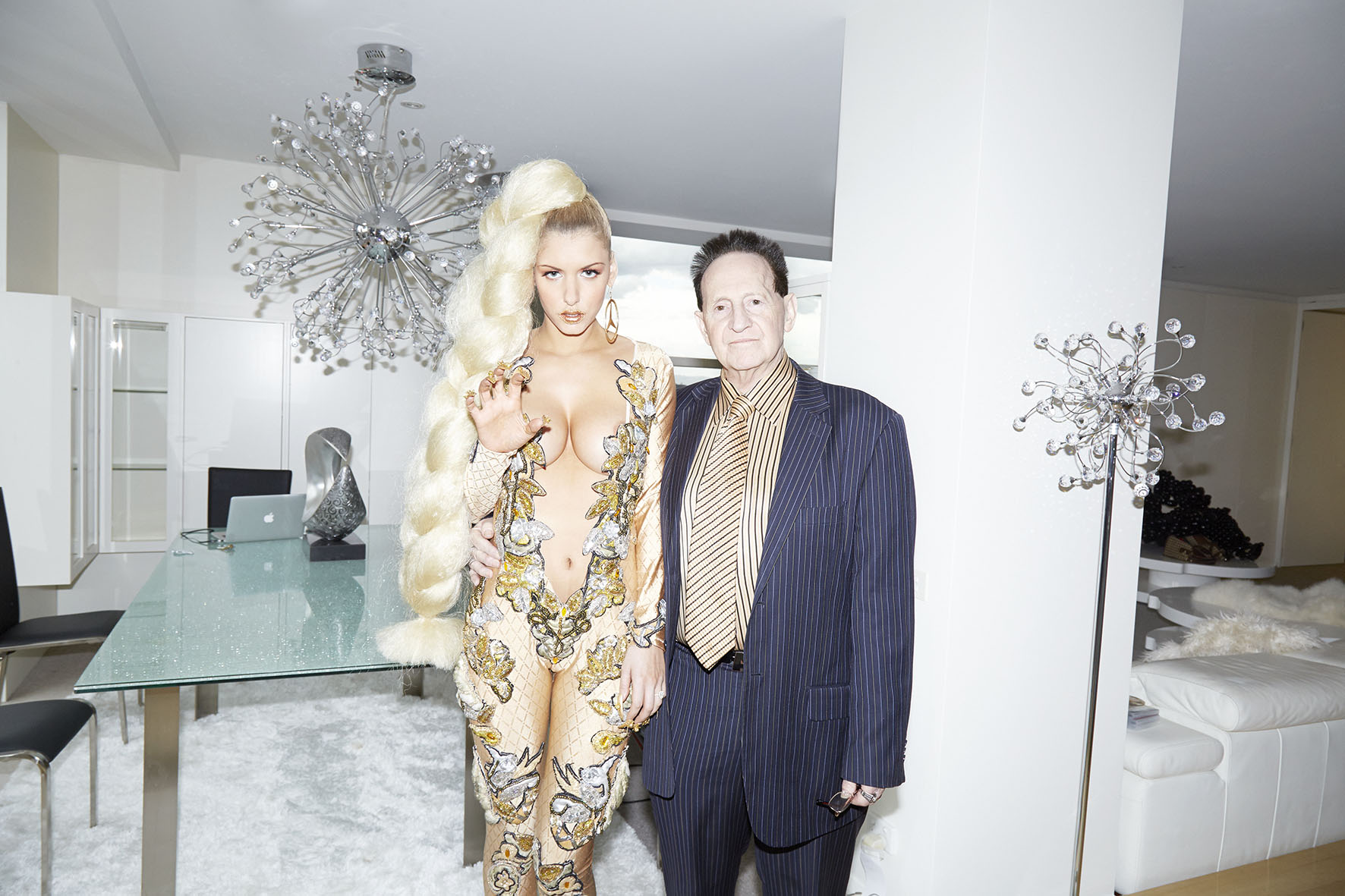 Blonde-haired woman in embellished bodysuit poses with an older man in pinstripe suit inside a modern apartment with chandelier.