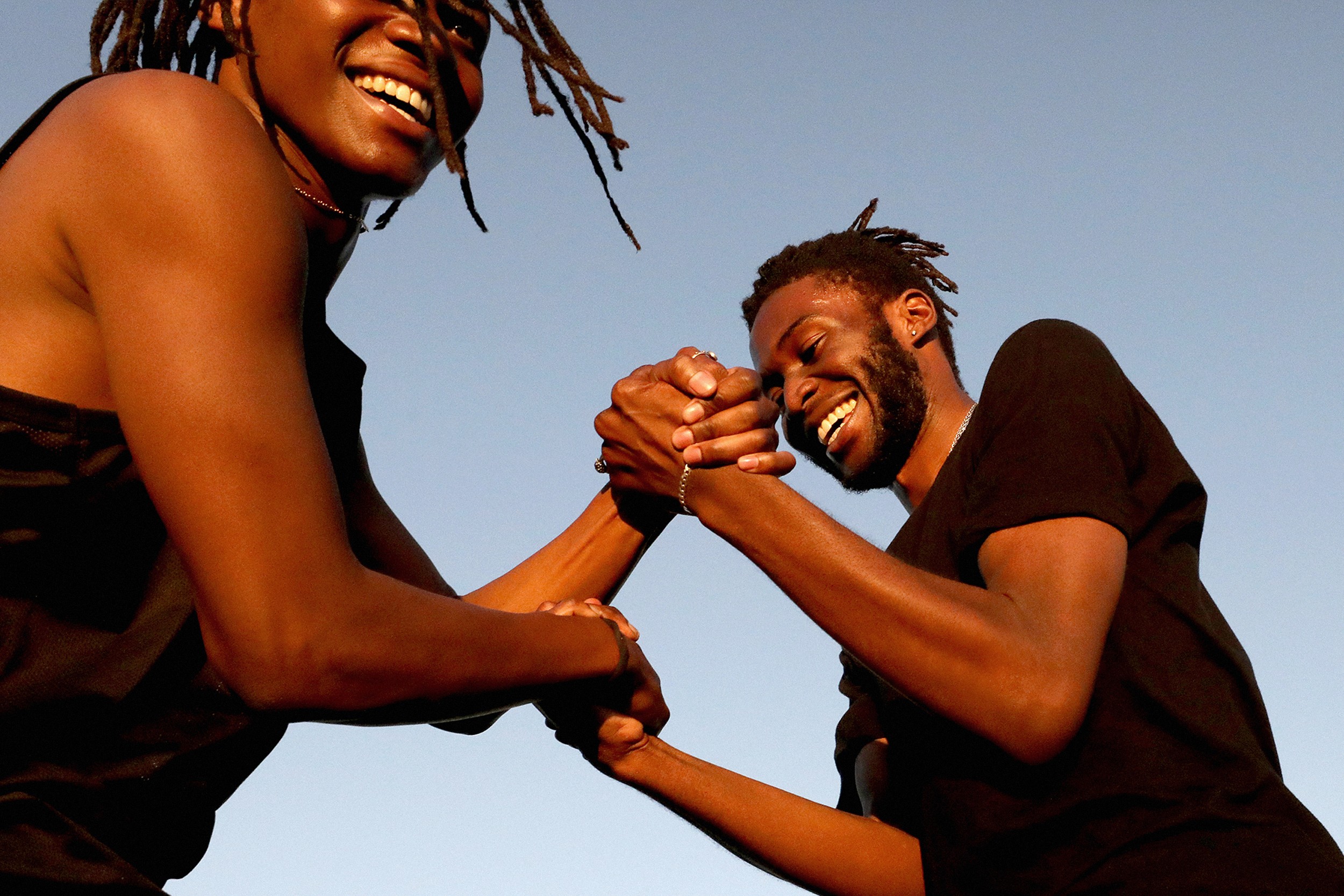 Two African American adults arm wrestling outdoors against a blue sky backdrop, displaying smiles and engaged in friendly competition.