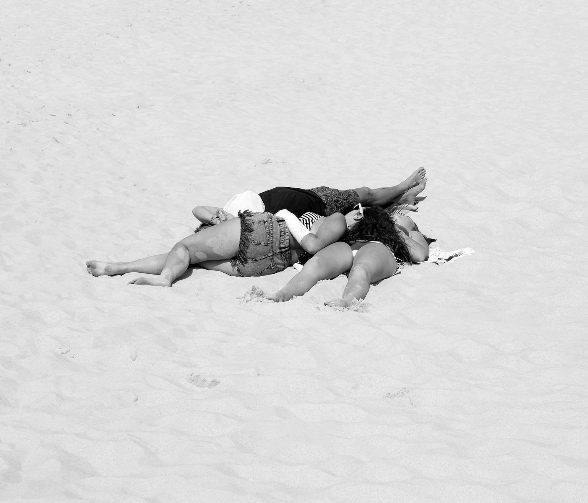 Three people rest on a sandy beach in a monochrome photograph. The group is close together, their bodies angled toward each other.