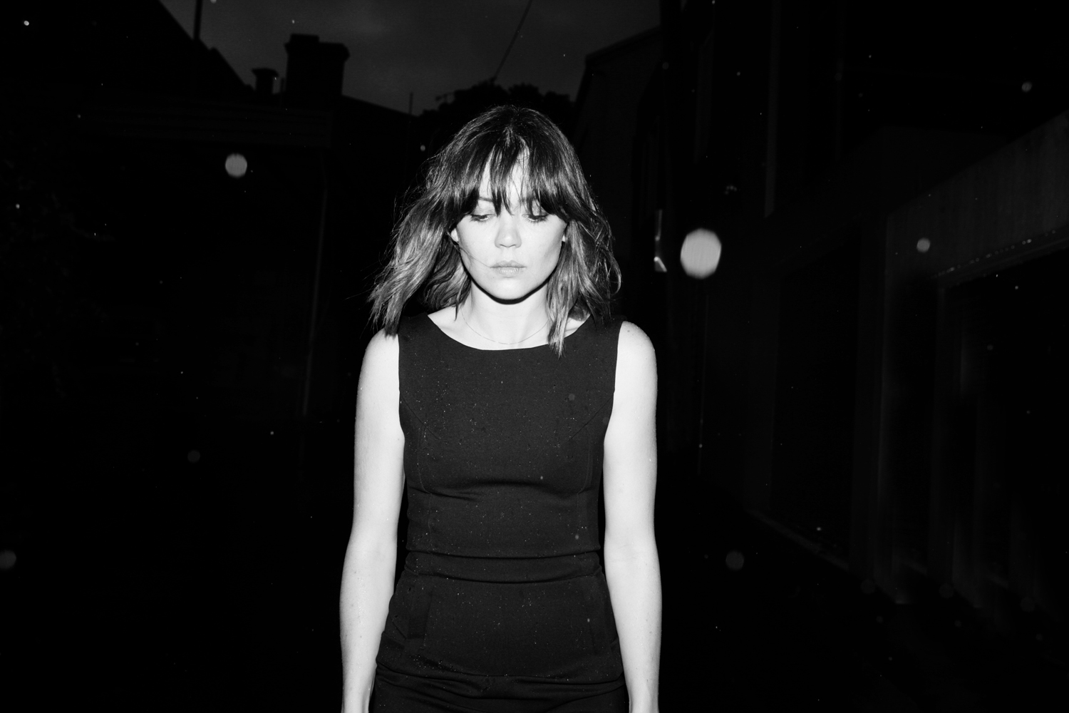 Monochrome portrait of a young woman with bangs and a dark sleeveless top, set against a dimly lit urban background. The photograph features a grainy texture.