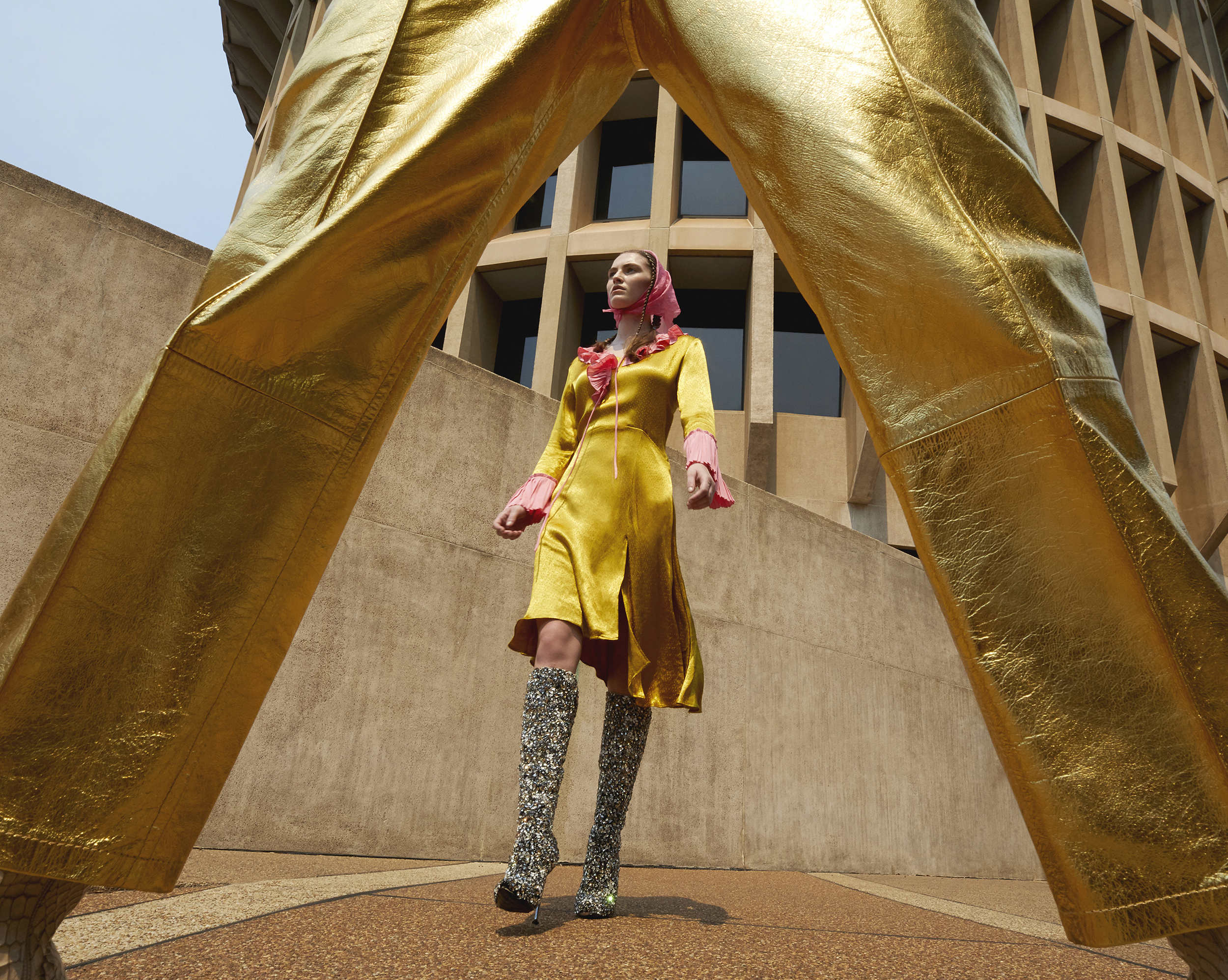 A woman in a yellow satin dress and patterned boots poses in front of a building. The frame is partially obscured by gold pants.