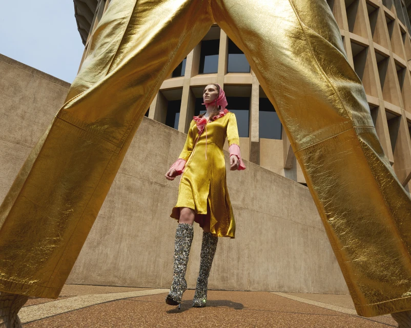A woman in a yellow satin dress and patterned boots poses in front of a building. The frame is partially obscured by gold pants.