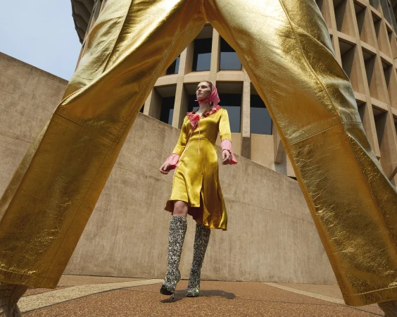 A woman in a yellow satin dress and patterned boots poses in front of a building. The frame is partially obscured by gold pants.