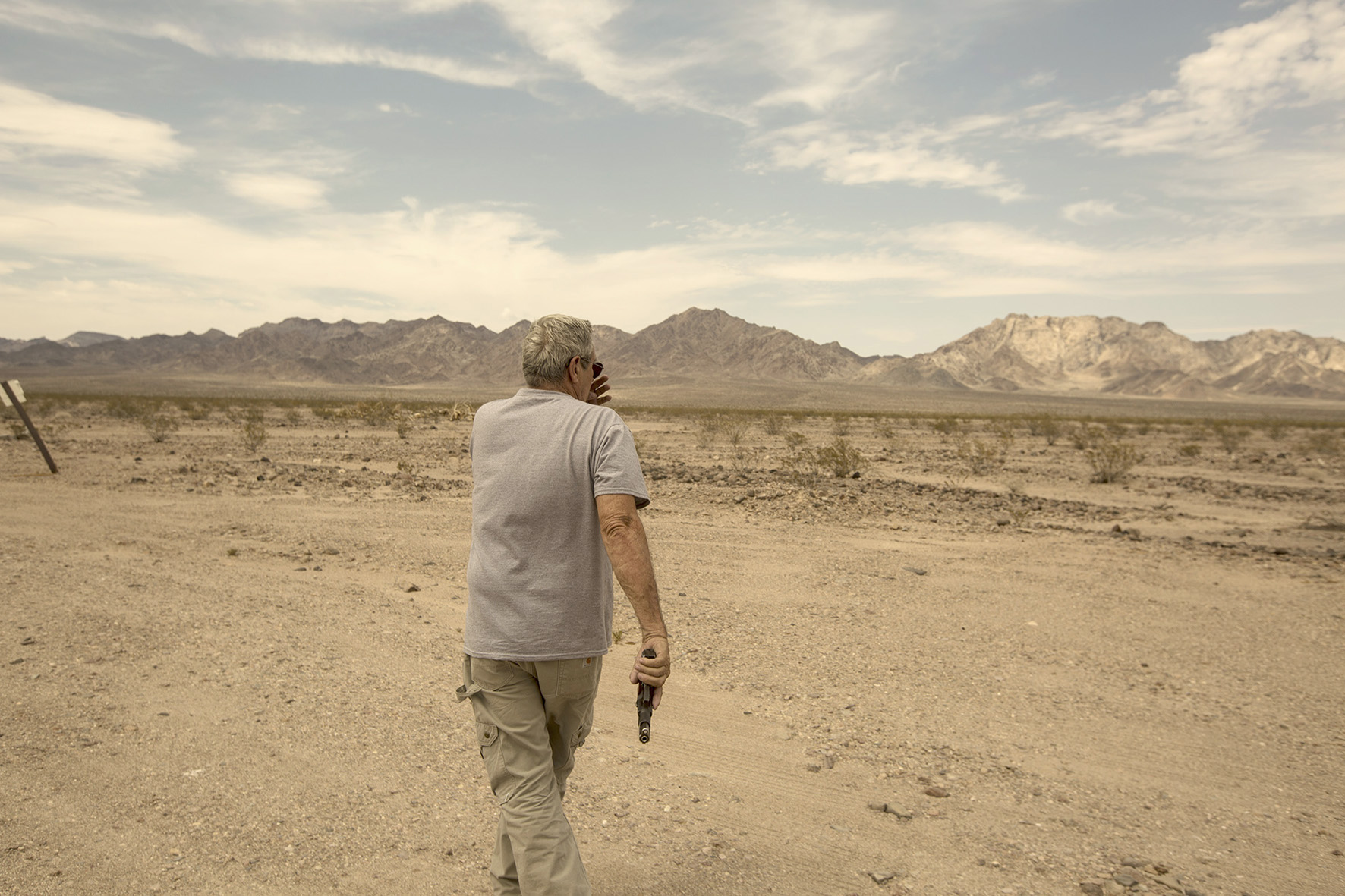 Man in casual clothing walks in the desert, holding a handgun, with a backdrop of arid mountains under a cloudy sky.