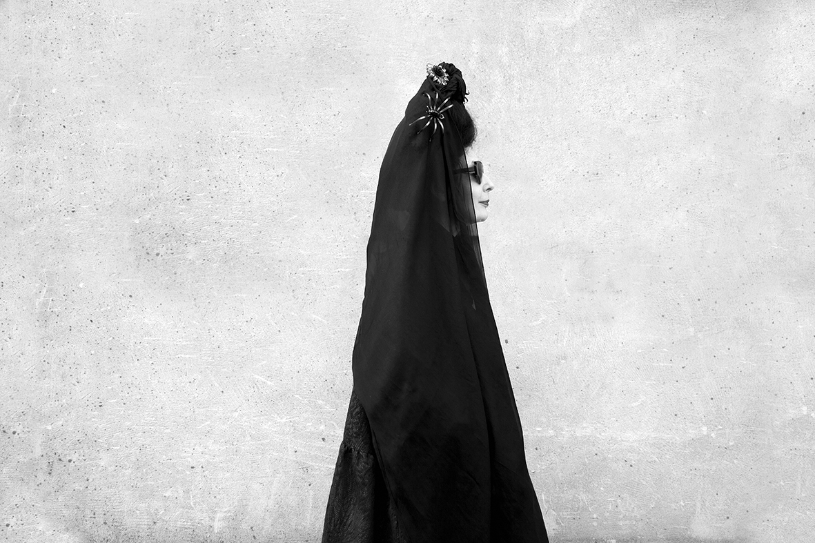 Woman in black dress and veil stands in profile against a mottled gray wall in a black and white photograph showcasing fashion and style.