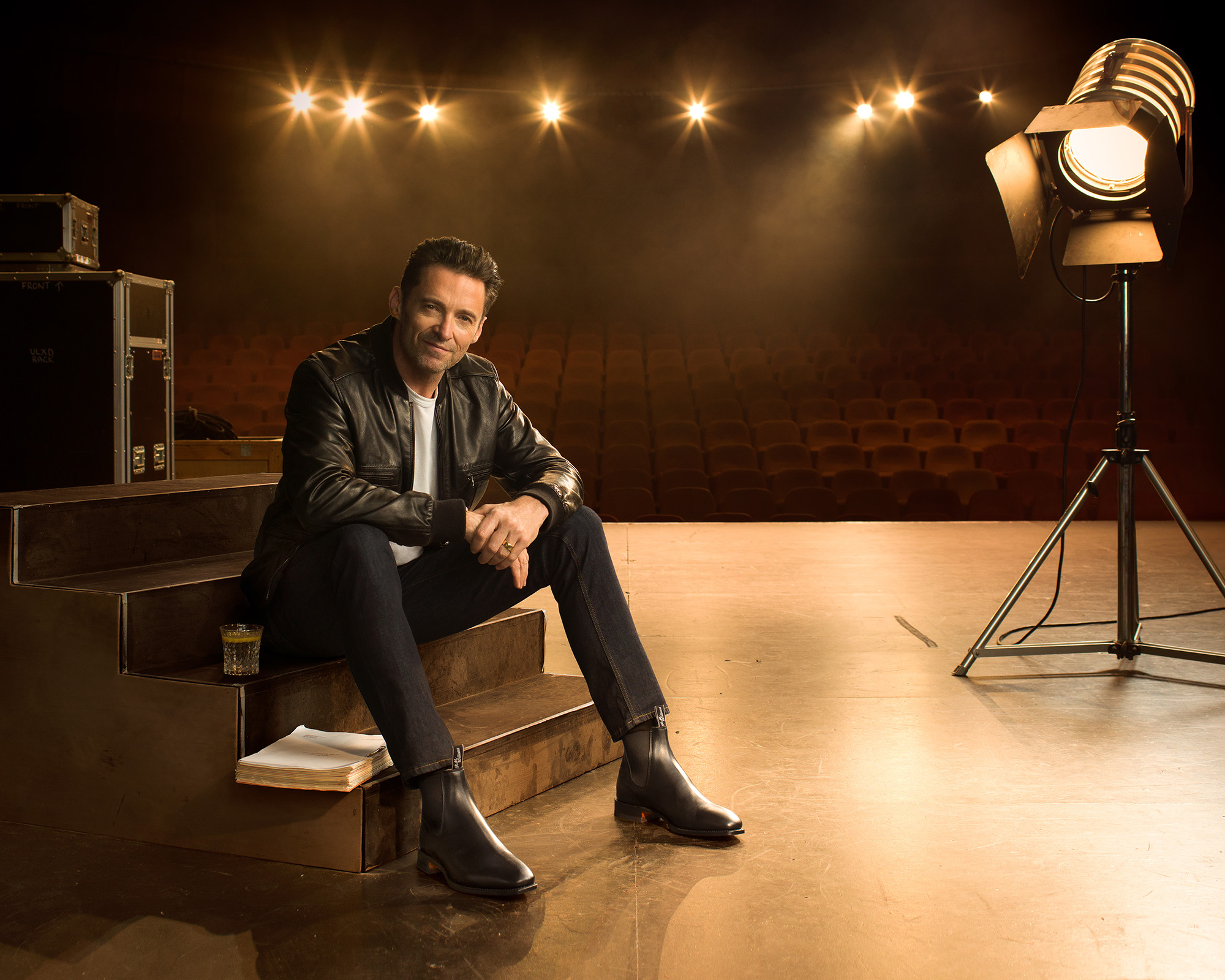 Hugh Jackman sits on stage stairs with a script and drink, lit by a stage light with rows of empty theatre seats in the background.