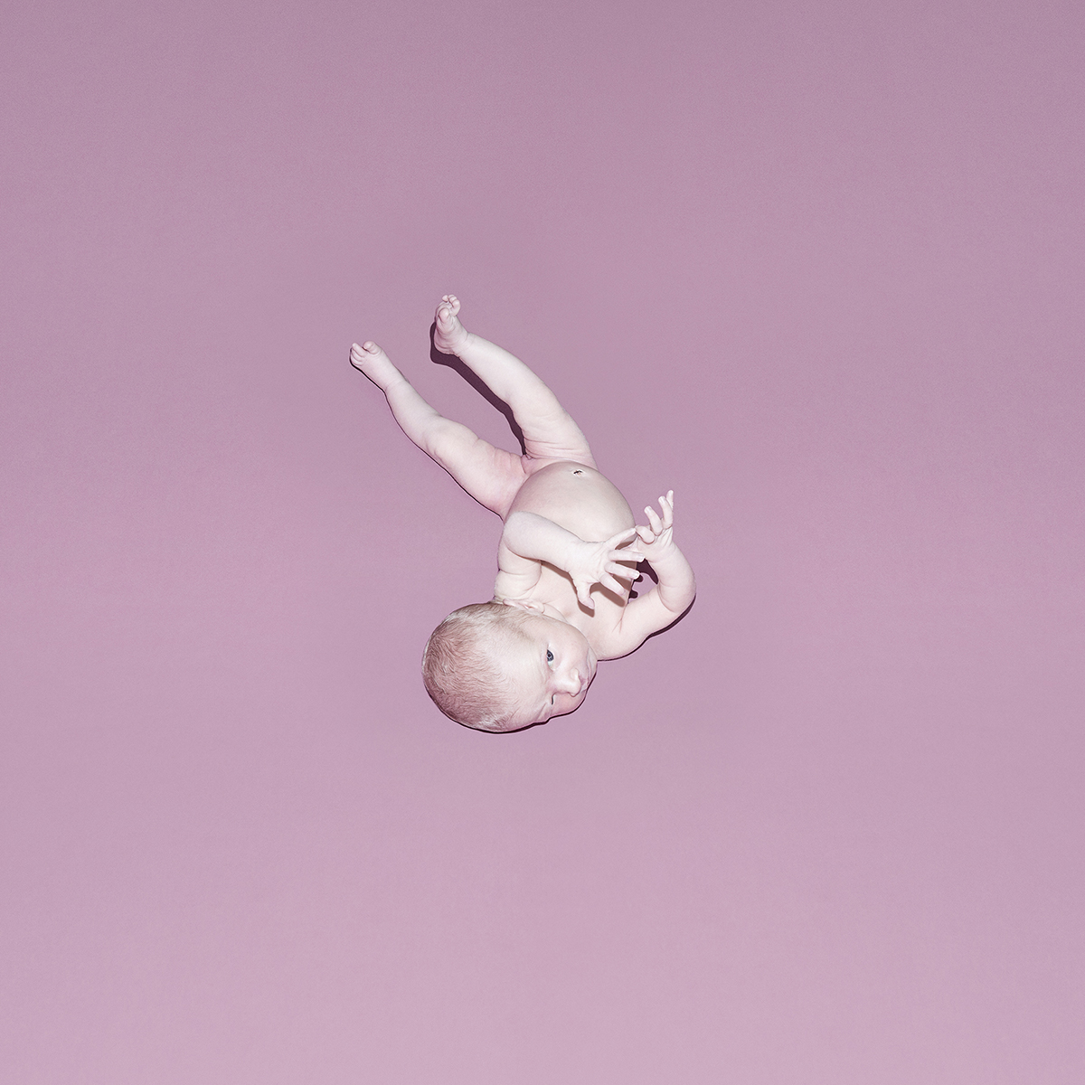 Overhead studio shot of baby doll on pink backdrop. The doll is positioned upside down, with hands near its head and feet elevated.