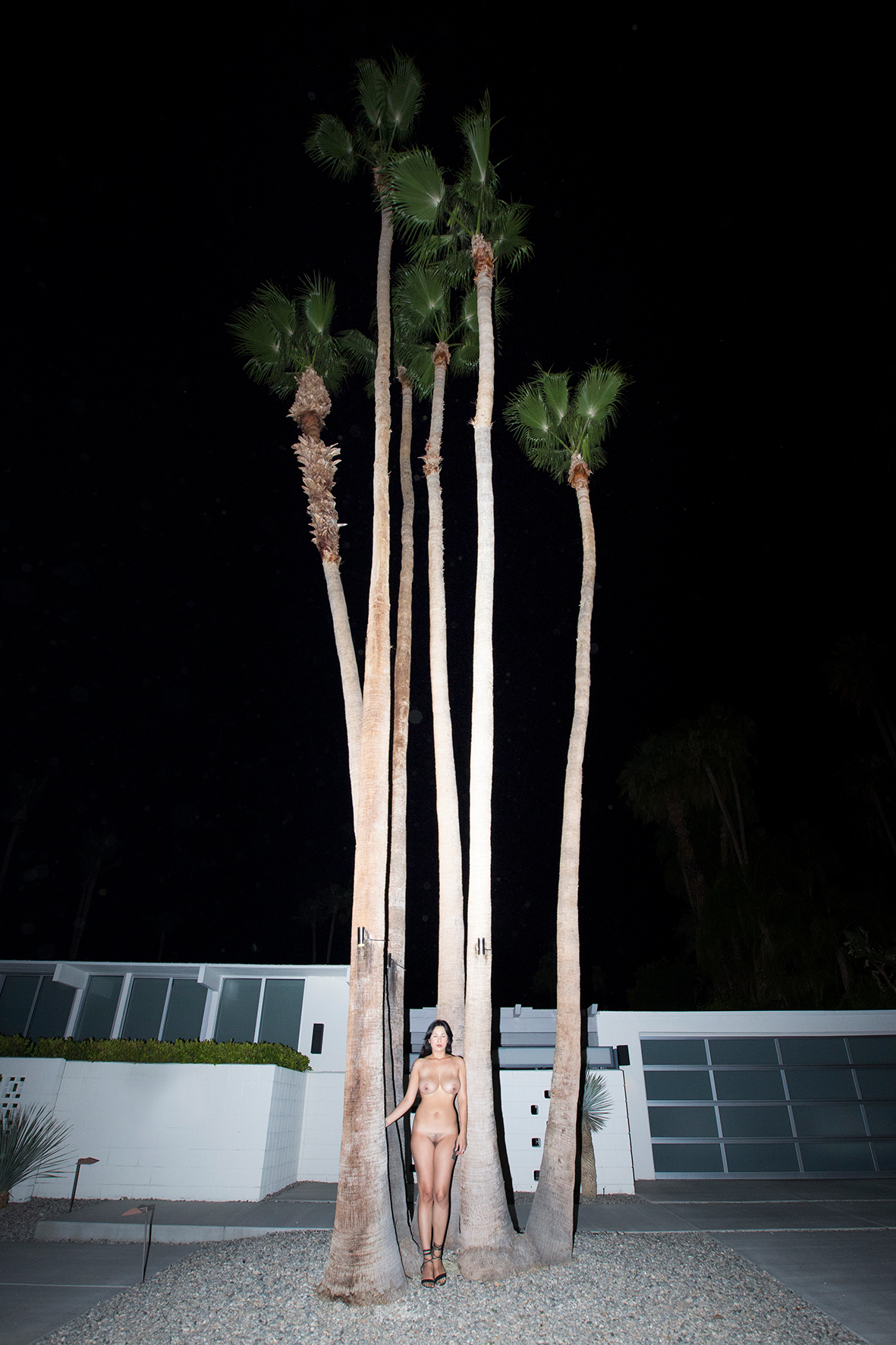 Woman poses next to palm trees in front of a modern building at night. Tall palms, dark sky, and residential architecture.