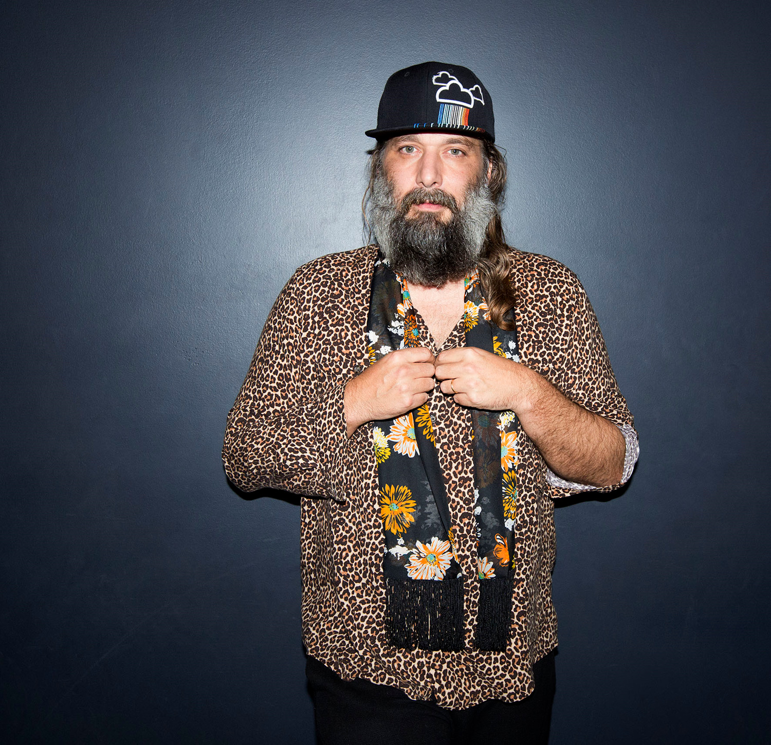 Adult male with a beard and patterned shirt posing indoors against a dark gray wall, wearing a ballcap and a floral scarf.