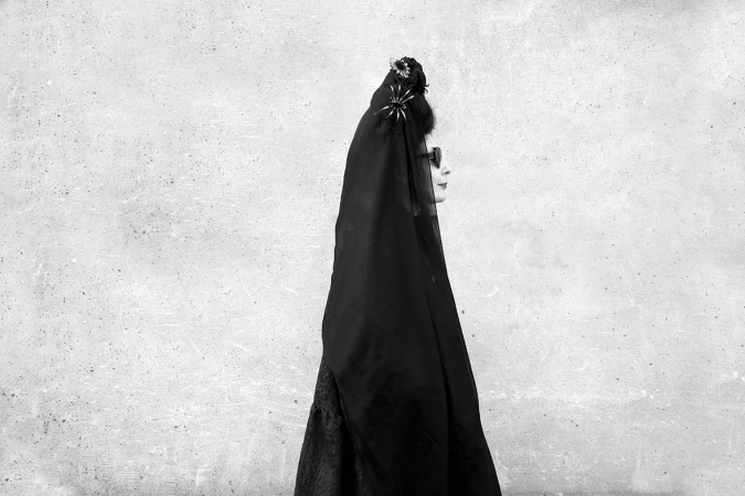 Profile portrait of a woman wearing a black veil and dark sunglasses against a textured concrete wall.