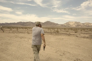 An older man walking away across a dry desert landscape holding a handgun, photographed by Juliet Taylor in Sydney.
