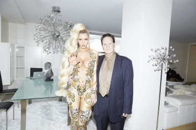Woman with very long blonde braid standing beside an older man in a pinstripe suit inside a modern living space.