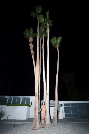A woman standing between tall palm trees at night in front of a modern house exterior.
