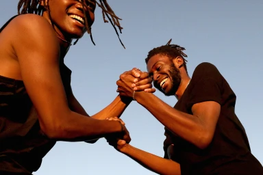 Two black men with dreadlocks smiling while arm wrestling against a clear blue sky
