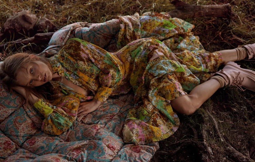 A woman with long hair lies on a patterned quilt, wearing a colorful floral print outfit and light pink boots, in a grassy outdoor setting.