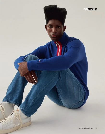 A young adult male model with a distinctive flattop hairstyle poses indoors, wearing a blue zip-up sweater, jeans, and white sneakers on a light background.
