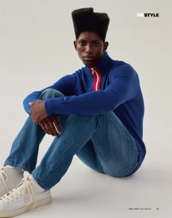 A young adult male model with a distinctive flattop hairstyle poses indoors, wearing a blue zip-up sweater, jeans, and white sneakers on a light background.