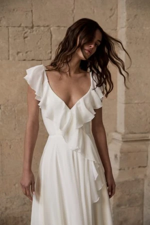 Woman with long dark hair wearing a white ruffled dress standing against rustic stone walls