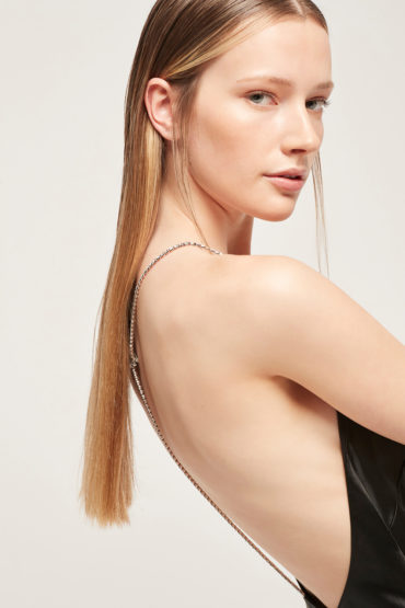 Close-up of fair-skinned adult woman, side view, with blonde hair wearing open-backed black dress and chain straps against neutral background.