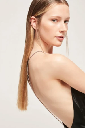 Close-up of fair-skinned adult woman, side view, with blonde hair wearing open-backed black dress and chain straps against neutral background.
