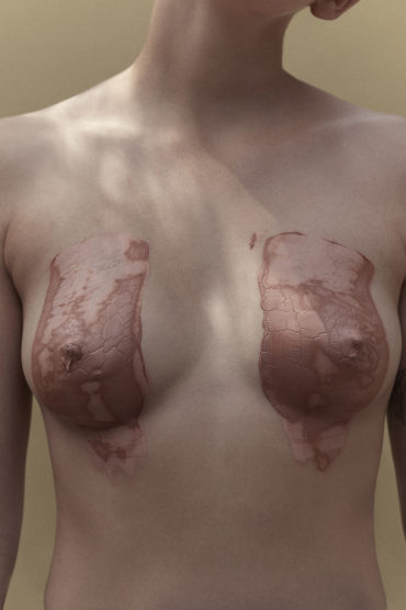 Artistic studio shot of a fair-skinned adult female torso with skin-tone body paint covering the breasts against a neutral background.