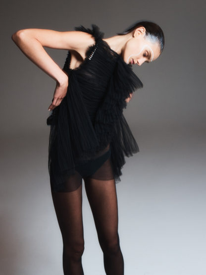 A fashionable woman in a black, frilled dress and sheer tights poses in a photo studio with a smooth gradient grey background.