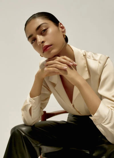 Elegant woman with slicked-back hair, wearing a silky shirt and leather pants, sits in a studio against a white background in a medium shot.