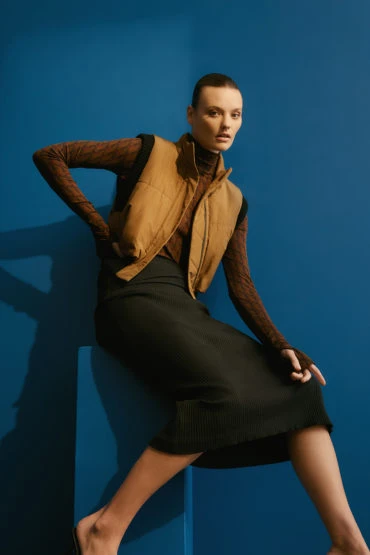 A woman in a brown top and vest, and black skirt, sits on a blue block against a blue background in a fashion studio shot.