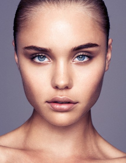 Close-up studio portrait of a young adult female with fair skin, light blue eyes, and light brown hair against a soft grey backdrop.