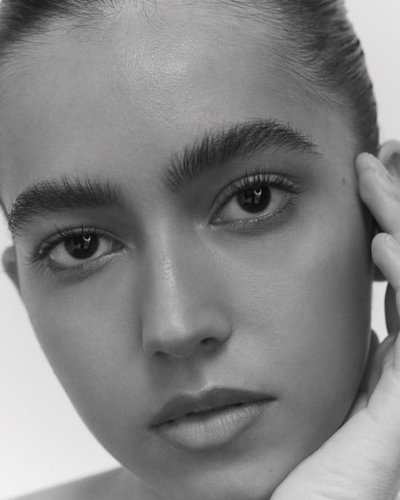 Close-up monochrome portrait of a woman with natural makeup, dark arched eyebrows, and hand resting near her face