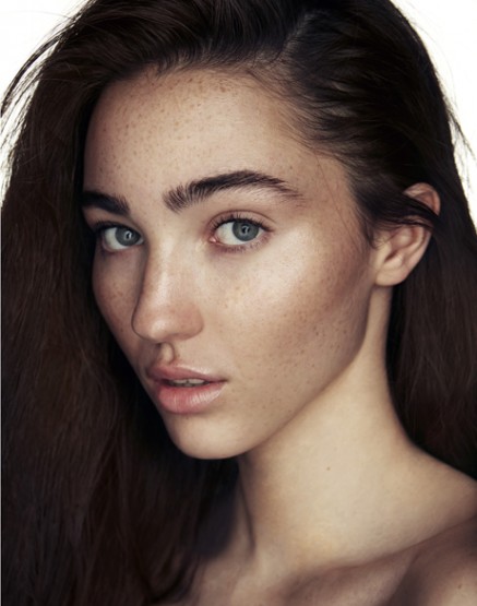 Close-up beauty portrait of a young woman with dark hair, green eyes, and visible freckles against a clean white background
