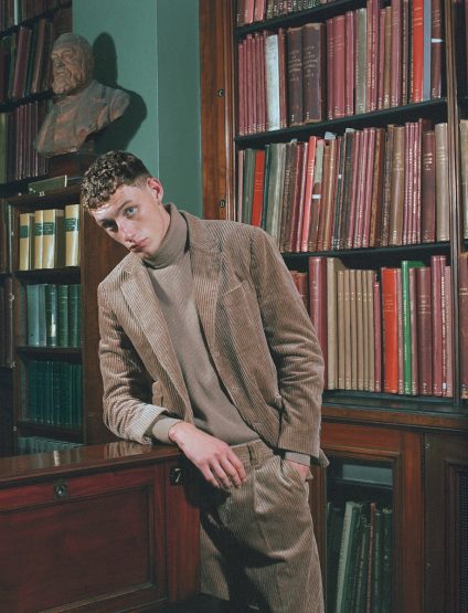 Adult male fashion model poses in a tan corduroy suit against a background of floor-to-ceiling bookshelves in a library interior.