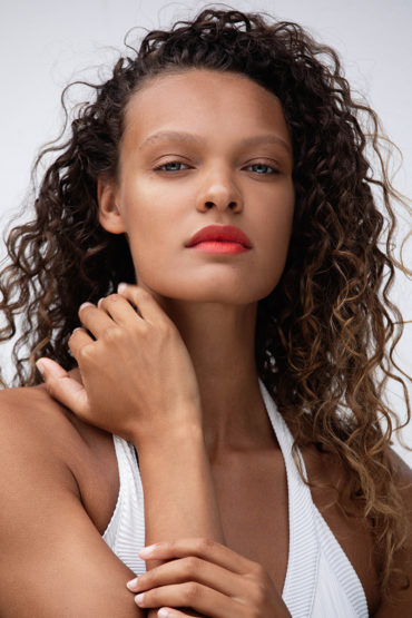 Portrait of a young woman with curly brown hair and coral lipstick, posing in a photo studio in a white ribbed tank top.