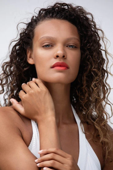 Portrait of a young woman with curly brown hair and coral lipstick, posing in a photo studio in a white ribbed tank top.