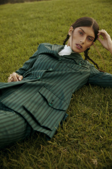 Model lying on grass wearing a green patterned jacket with braided hair and natural makeup by Joel Phillips