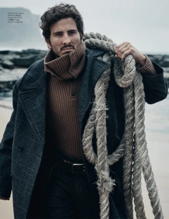 Man in dark wool coat and ribbed turtleneck sweater holding a thick rope on a beach with rocky cliffs in the background