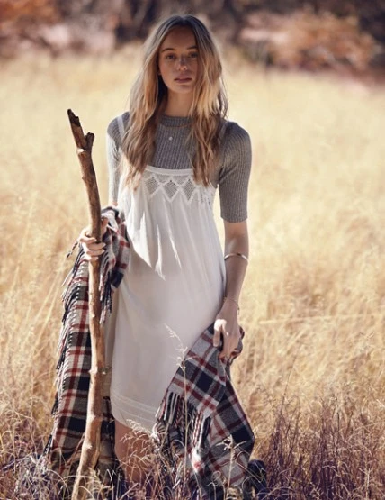 Young woman with long hair in a white dress and grey top holds a wooden stick with a plaid blanket in a grassy field setting.