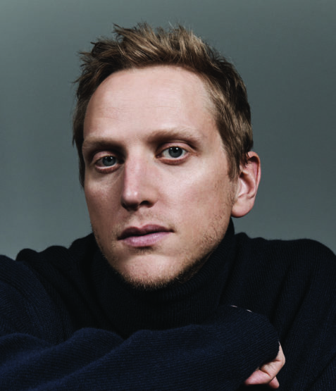 Close-up portrait of a man with light hair wearing a dark turtleneck against a plain dark grey background.