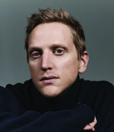 Close-up portrait of a man with light hair wearing a dark turtleneck against a plain dark grey background.