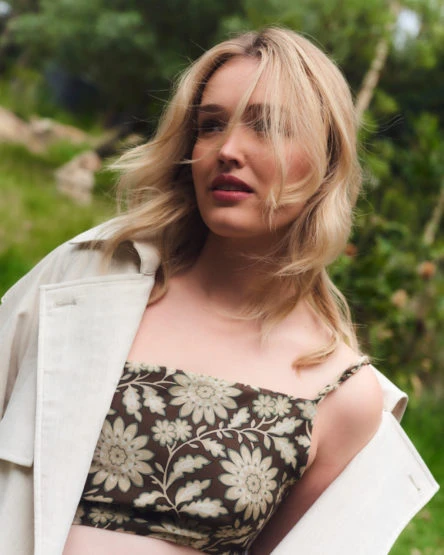 Blonde woman in a patterned crop top and jacket posing outdoors against a blurred green background, looking off-camera with windblown hair.