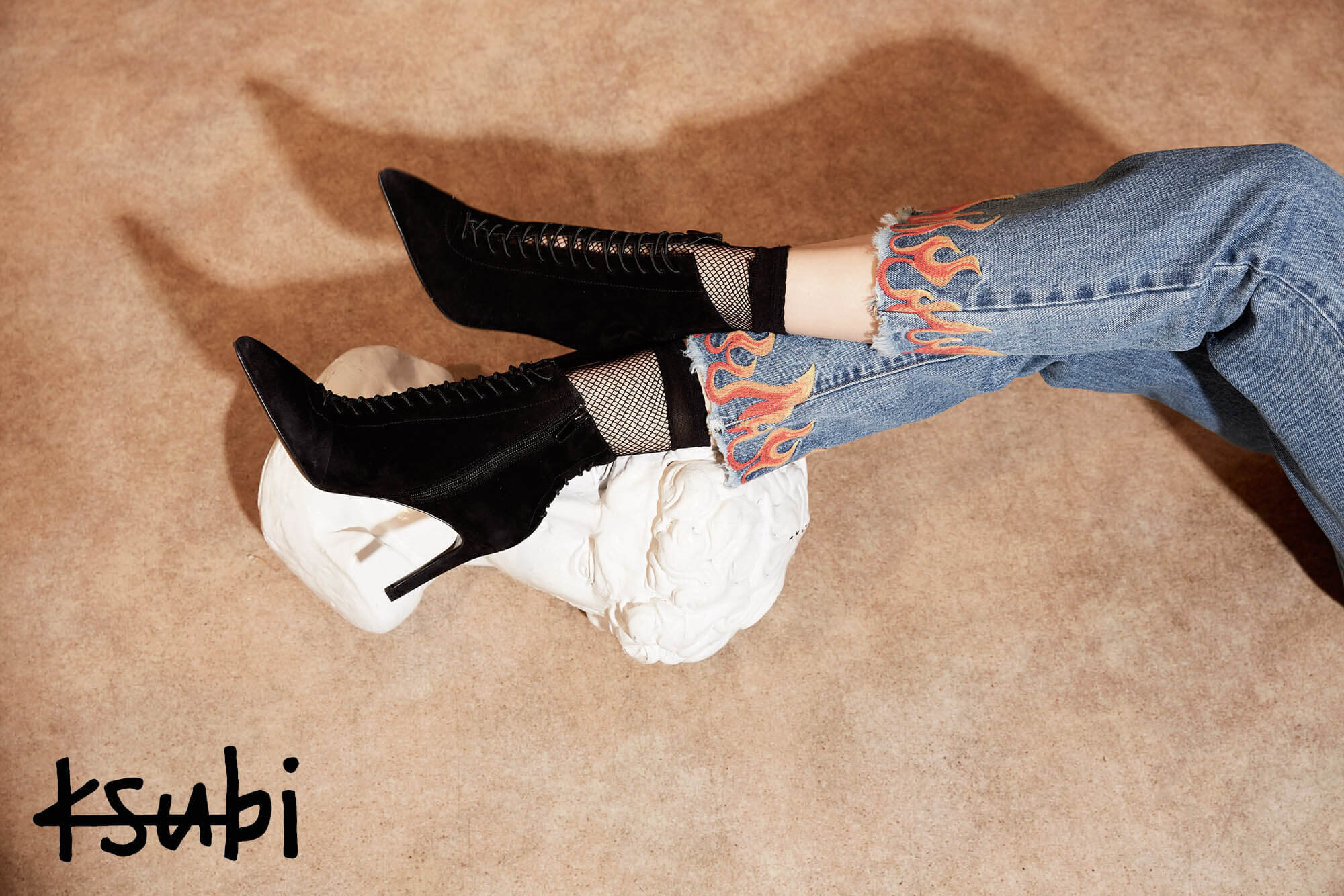 Black lace-up heeled boots rest on a white sculpture, paired with flame-design jeans against a neutral backdrop. Ksubi logo visible.