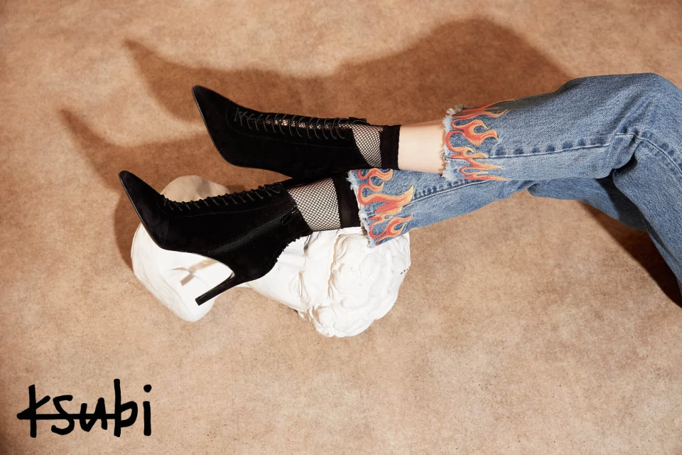 Black lace-up heeled boots rest on a white sculpture, paired with flame-design jeans against a neutral backdrop. Ksubi logo visible.