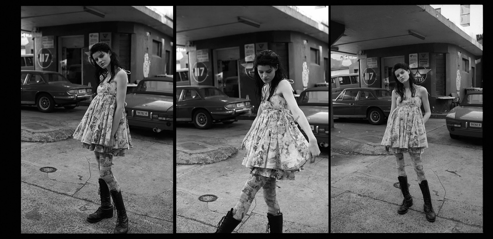 Monochrome image of a young woman in a dress and boots stands outside near vintage cars and a retro gas station building.