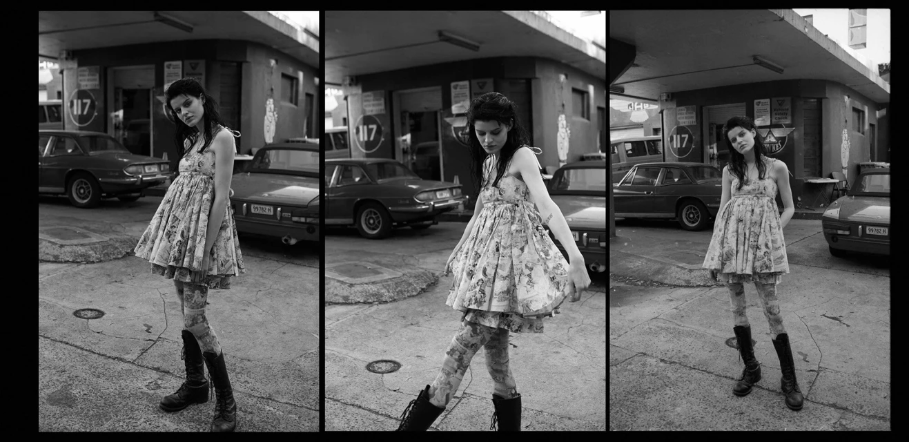 Monochrome image of a young woman in a dress and boots stands outside near vintage cars and a retro gas station building.